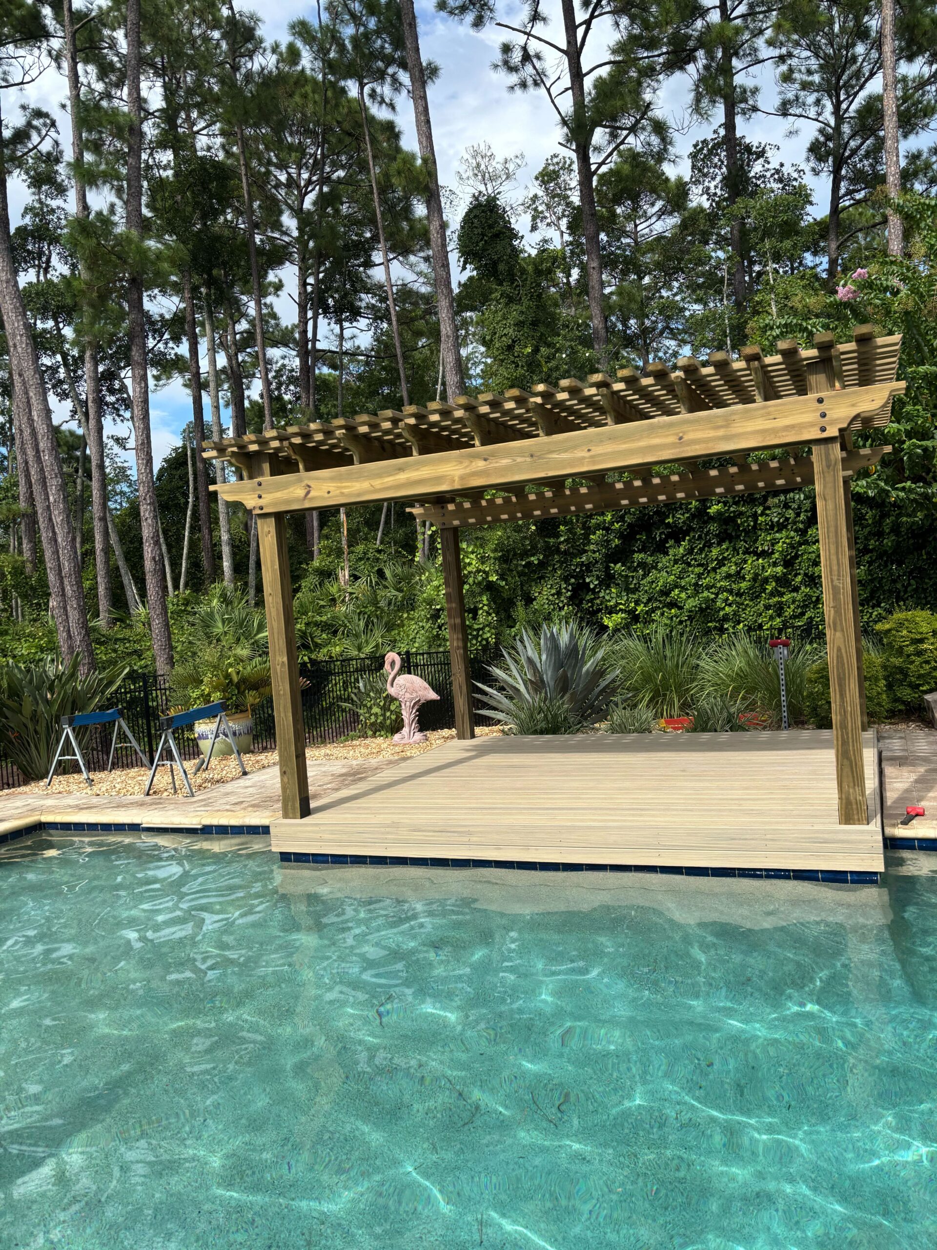 Wooden patio cabana structure beside a clear pool, surrounded by lush greenery and a pink flamingo statue, showcasing durable outdoor design by Coastal Outdoor Construction.