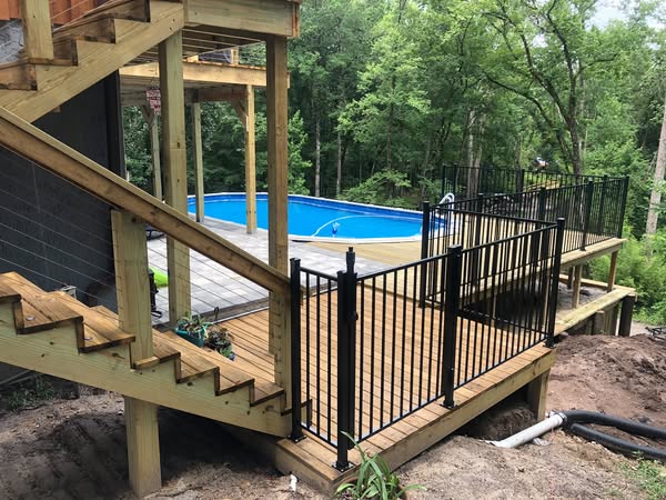A wooden deck with black railing and stairs, leading to an above-ground pool surrounded by lush greenery.