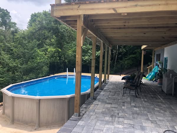 Composite pool deck with an above-ground pool, lush greenery, and a shaded patio with seating and a slide.