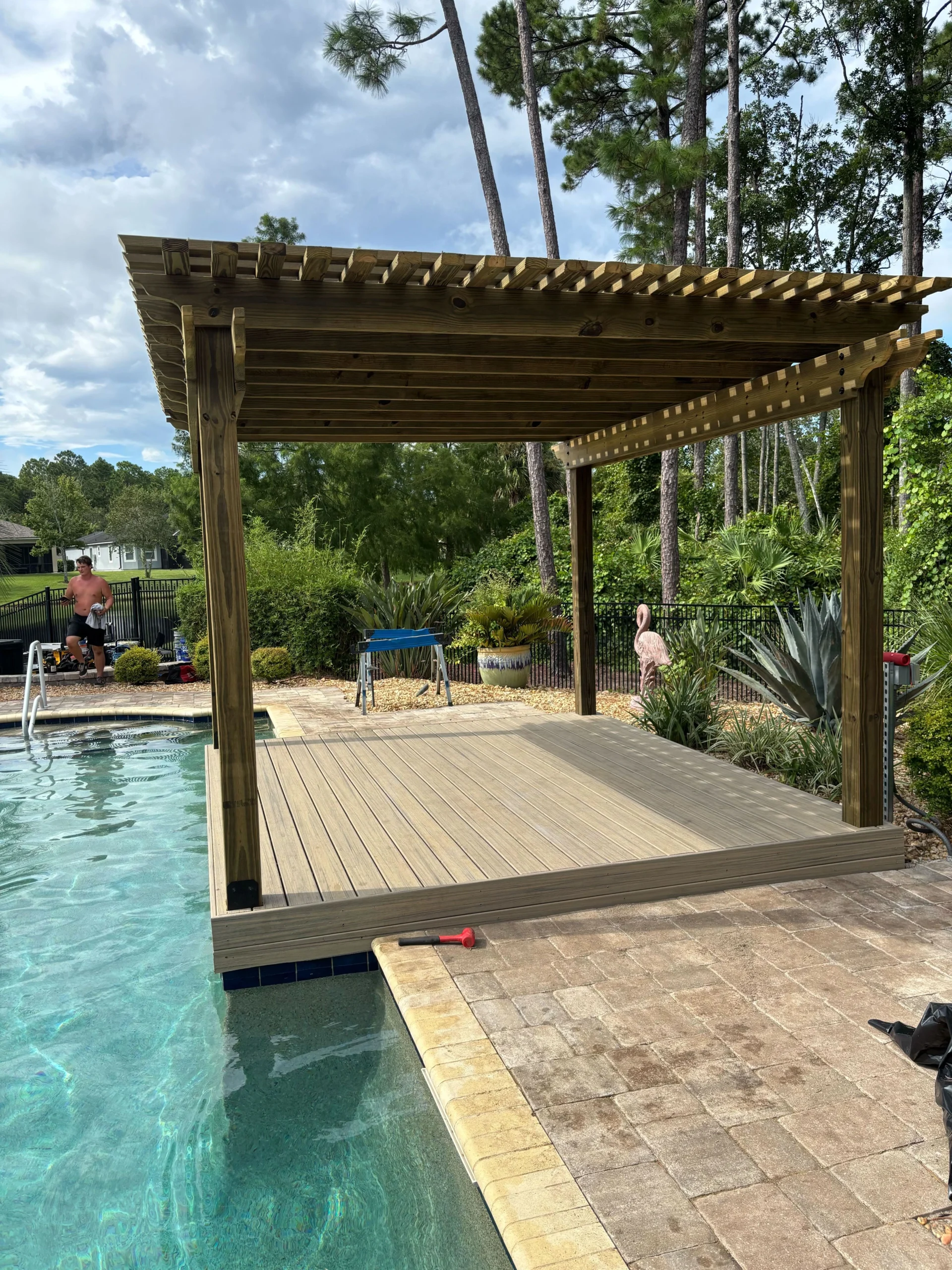 Wooden pergola structure beside a swimming pool, designed for outdoor relaxation, showcasing custom craftsmanship by Coastal Outdoor Construction, surrounded by lush greenery and patio space.